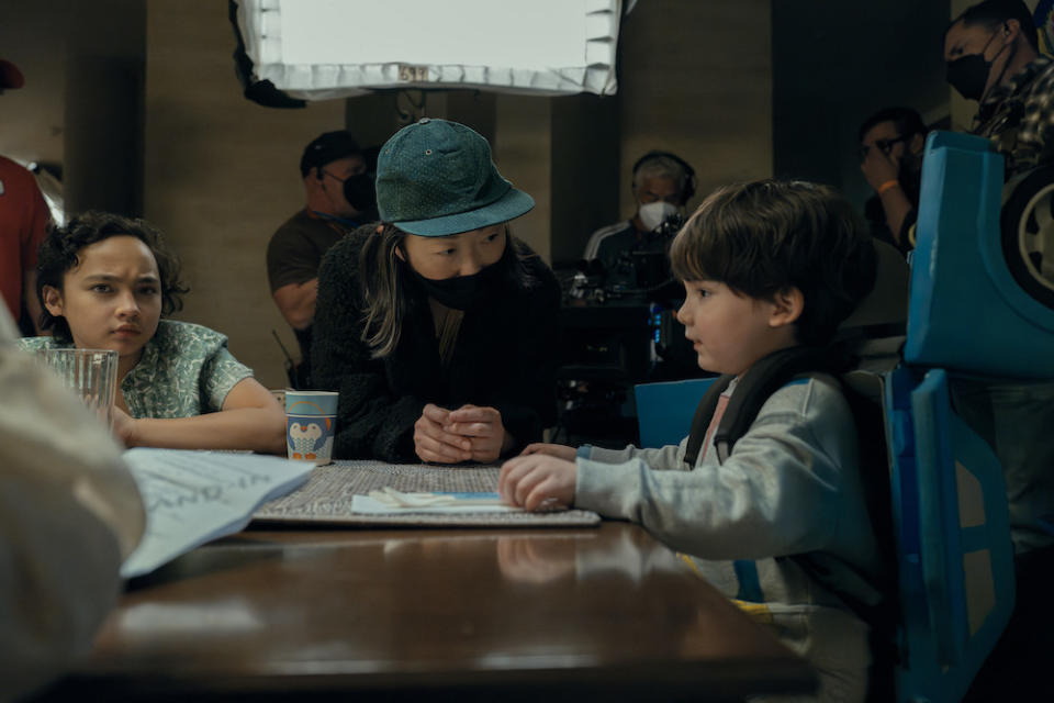 Lulu Wang, wearing a green baseball cap and a black facemask, hunches over a table to talk to two child actors on the set of 'Expats'
