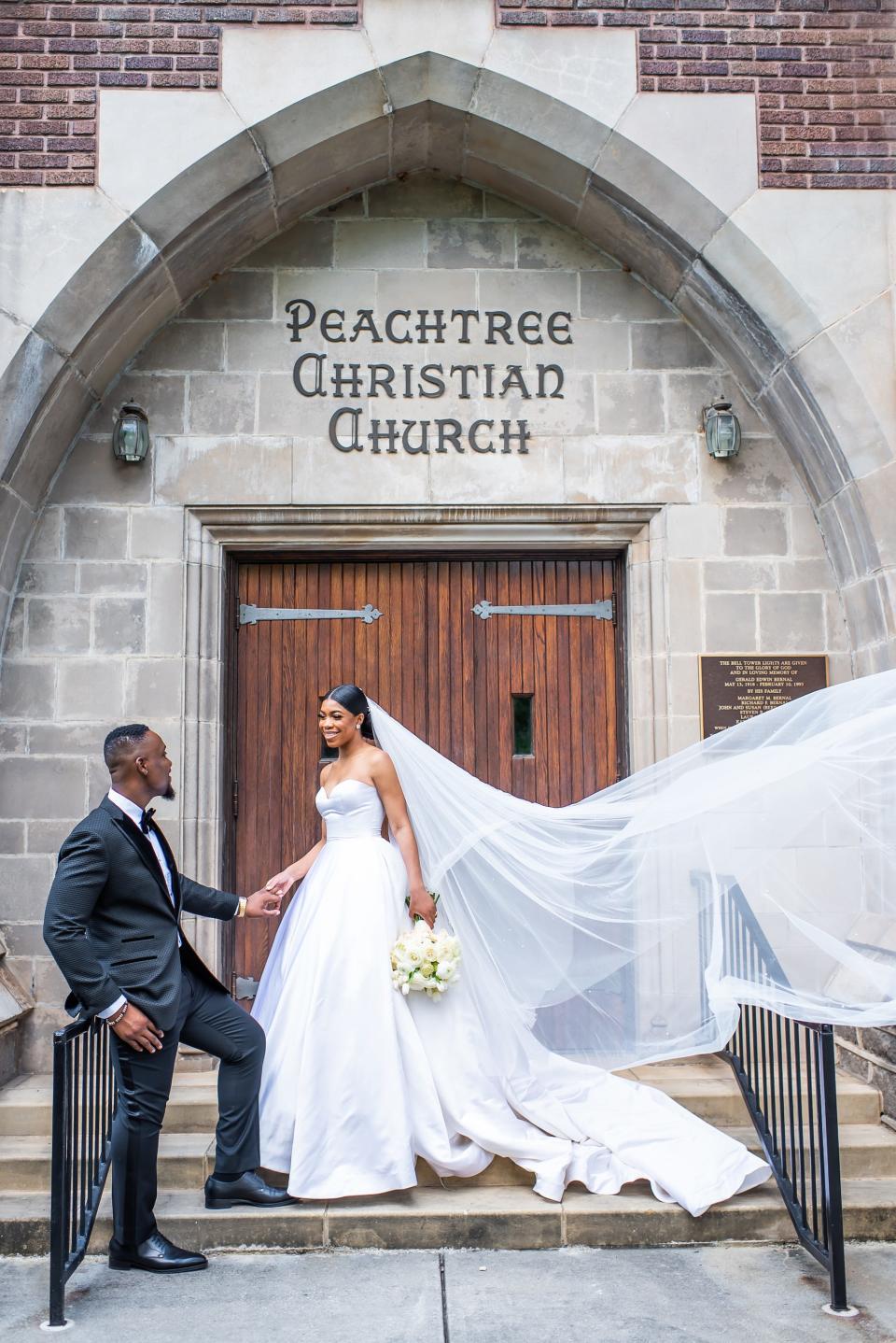 A bride's veil flies up as she and her groom look at each other in front of a church on their wedding day.