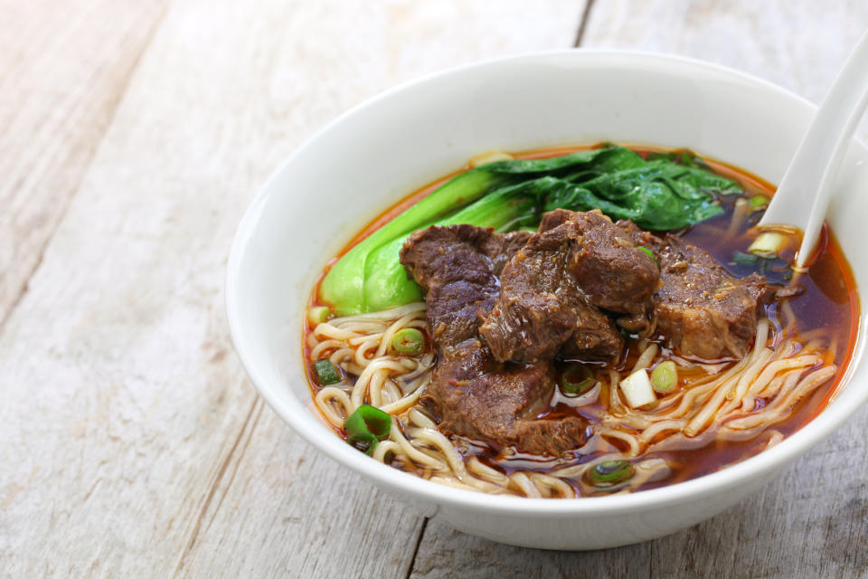 Bowl of beef noodle soup with bok choy, beef chunks, scallions, and a spoon on a wooden surface