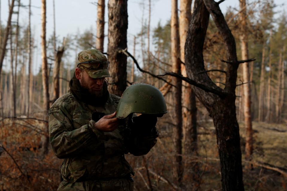 A man in camouflage stands in the woods holding and looking down at a helmet.