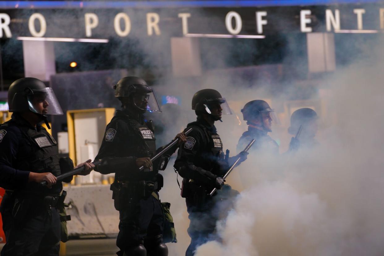 Agents in riot gear stand holding clubs amid smoke near a port of entry