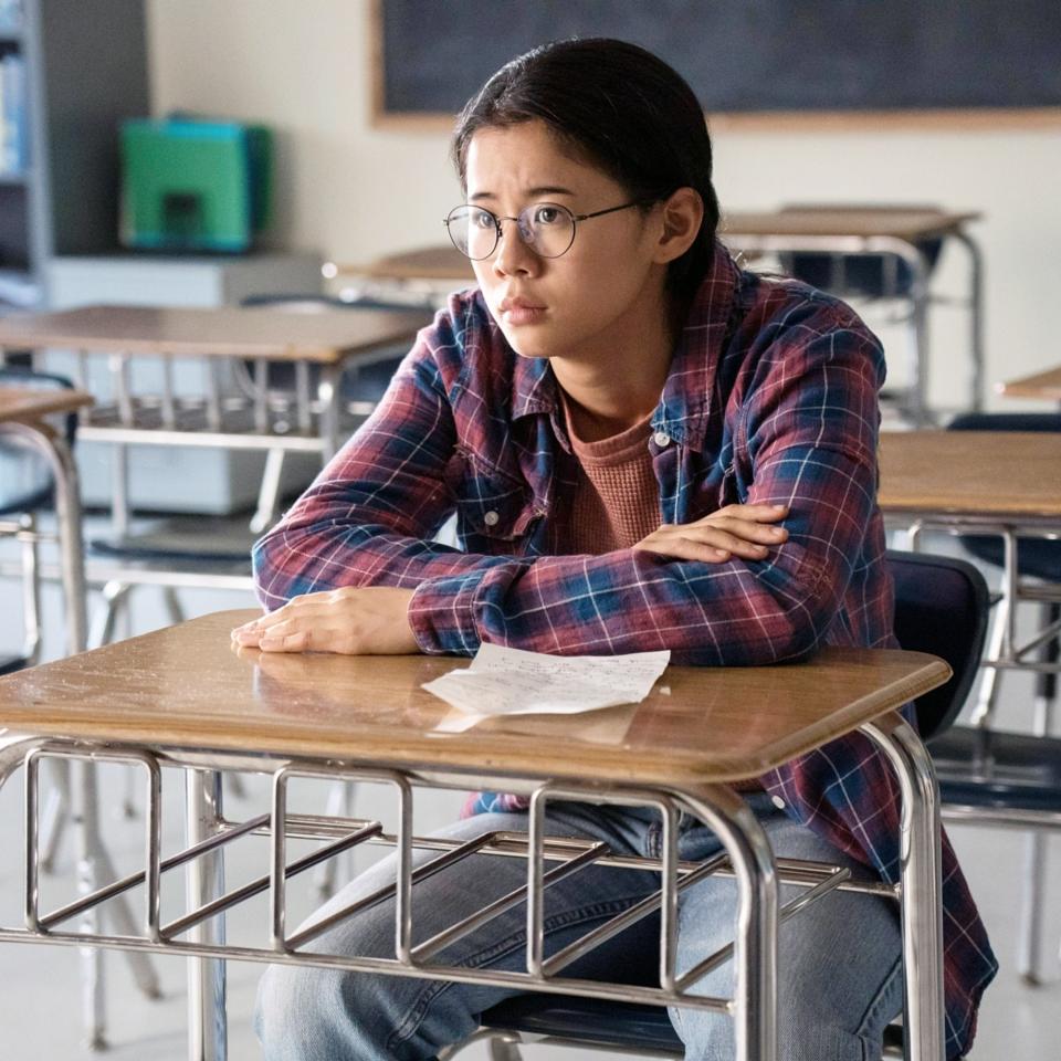 A person wearing glasses, a plaid shirt, and jeans sits at a wooden desk in an empty classroom, looking pensive with a note on the desk