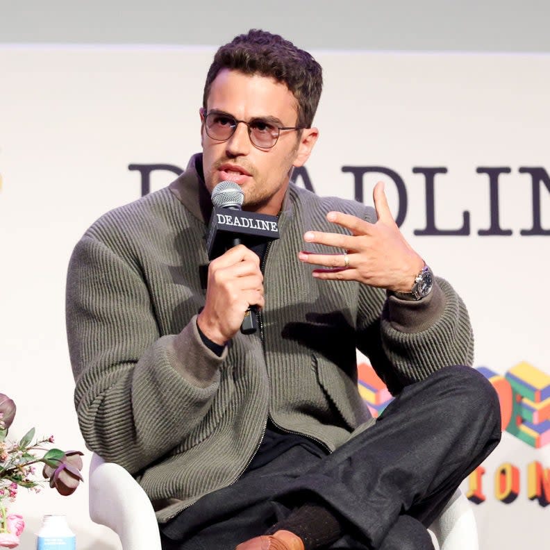 Theo James speaks into a microphone while sitting in a chair at a Deadline event, wearing glasses, a knit sweater, and dark pants