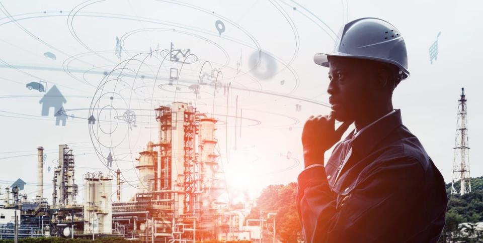 A person wearing a hard hat appears to be thinking while surrounded by various oil and gas infrastructure in the background.  