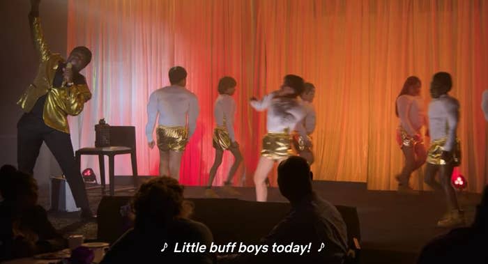 sam richardson wears a shiny suit and small afro wig. he sings into a mic, "little buff boys today!" while young adult boys wear fake muscle suits and shiny shorts.