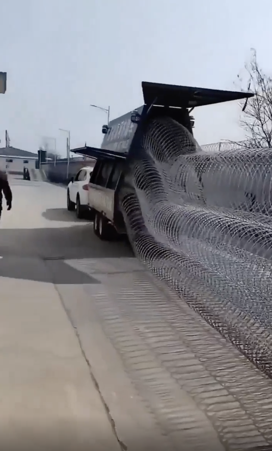 Truck bed overfilled with large rolls of wire mesh, some unspooling onto street, person nearby