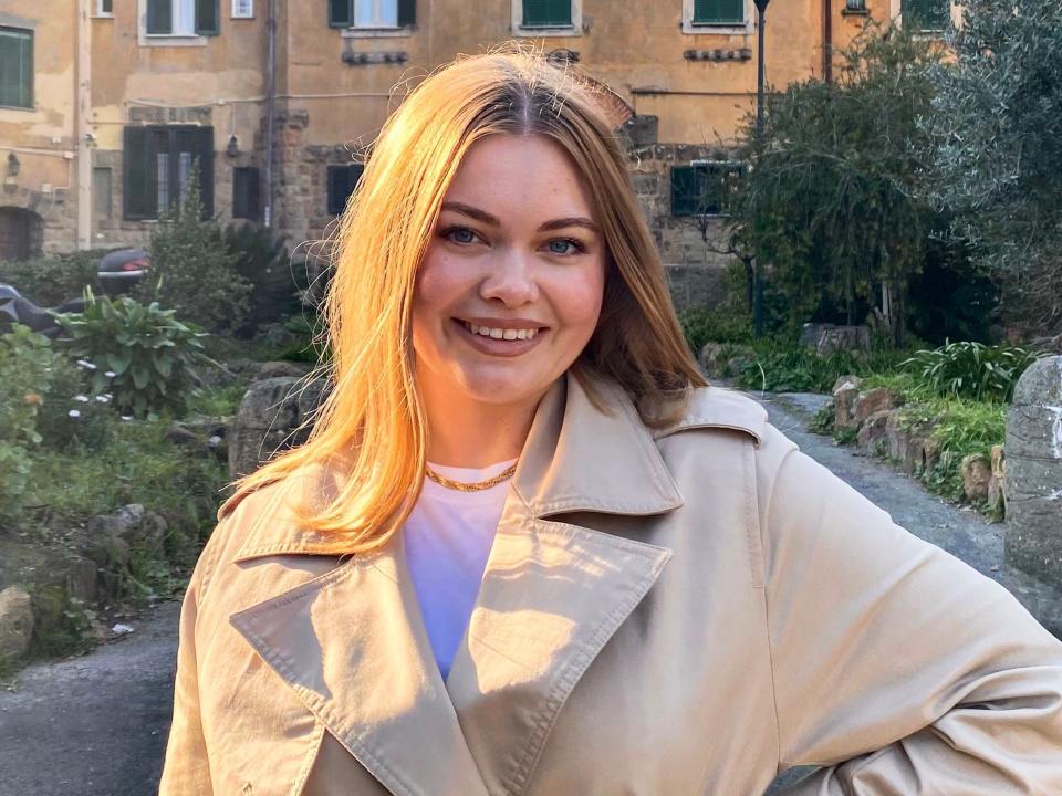 The writer, wearing a white shirt and trench coat, smiles at the camera with a neutral toned-lip color. She stands in front of a terrace and a residential building in Italy