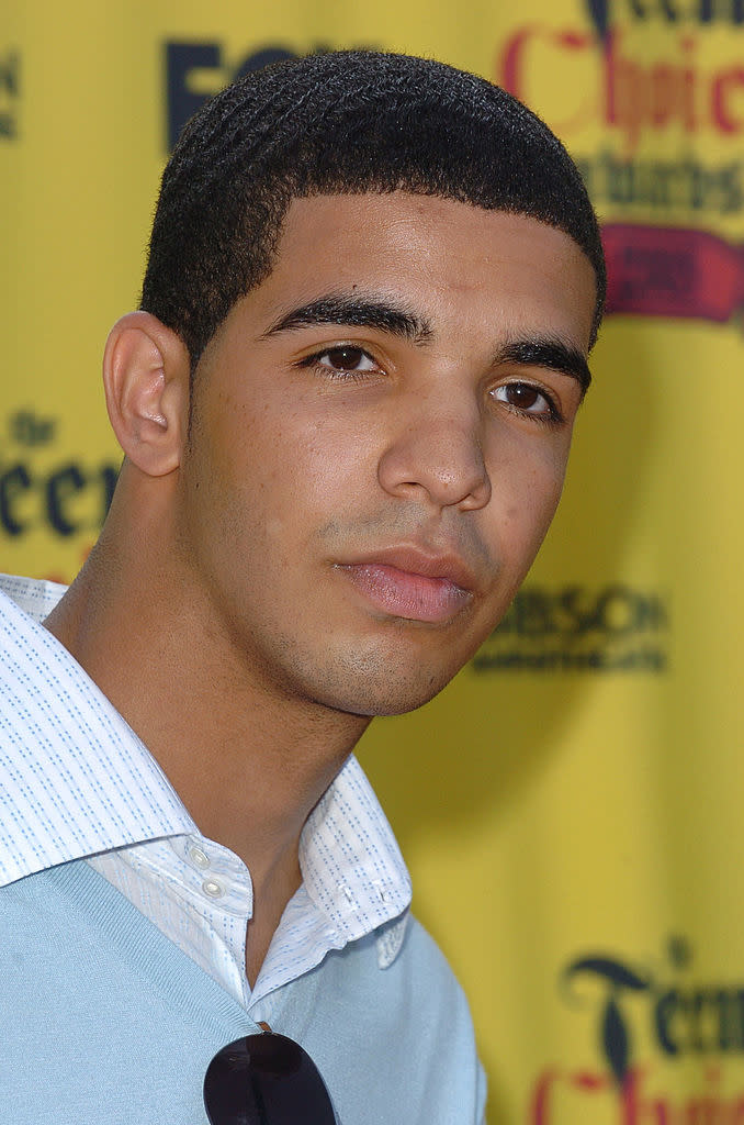 Drake at a red carpet event, wearing a light blue sweater over a white collared shirt, with sunglasses hanging from his sweater