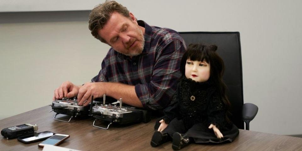 Animatronics designer Paul Jones sits at a table with the Nadja doll sitting on top of the table. Jones is looking at the doll as he adjusts a remote controller to move its head and eyes. 