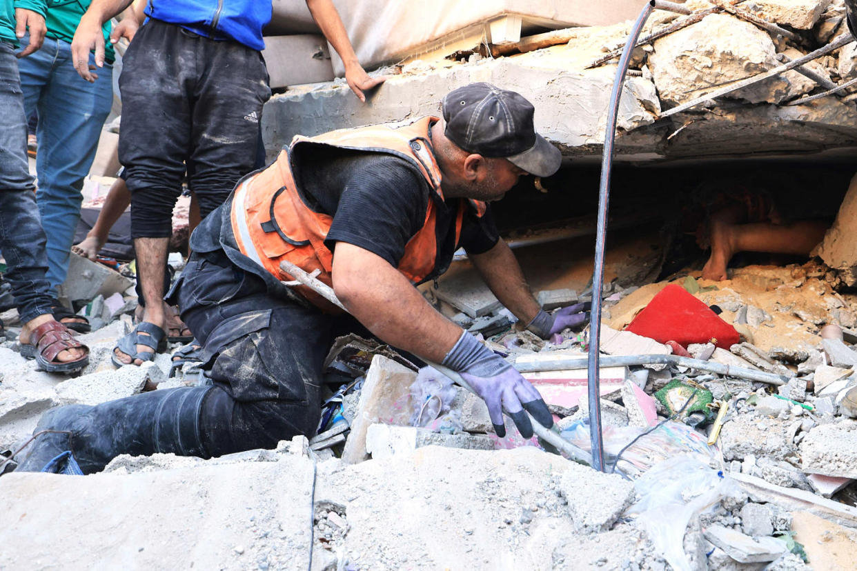 A man in an orange emergency vest kneels and speaks to a person underneath building rubble ( Mahmud Hams / AFP via Getty Images)