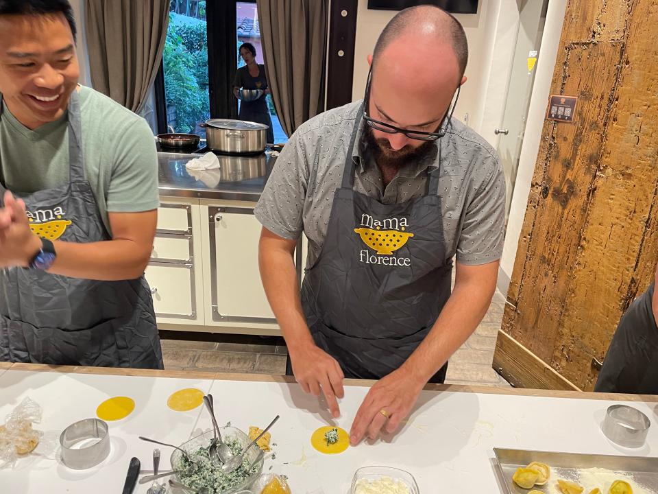 A man wearing a gray shirt and an apron with "mama florence" text and a graphic of a yellow colander makes circular ravioli