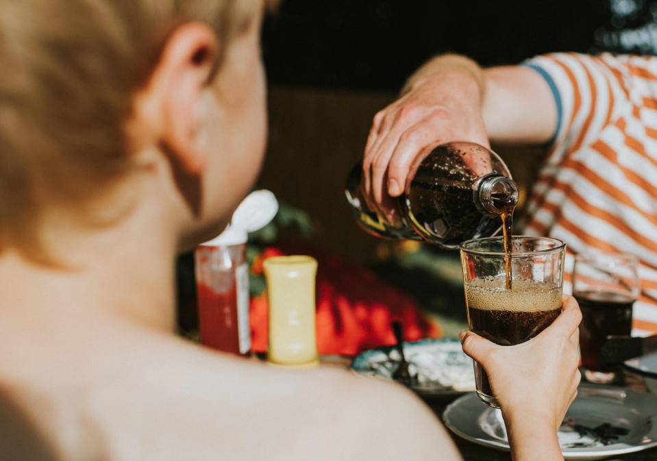 Person pouring a dark beverage into a glass at an outdoor gathering with others around a table