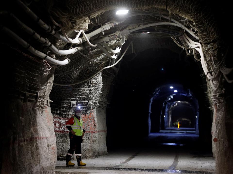 worker in neon green vest hardhat and respirator mask stands at the side of a long tall dark tunnel deep underground