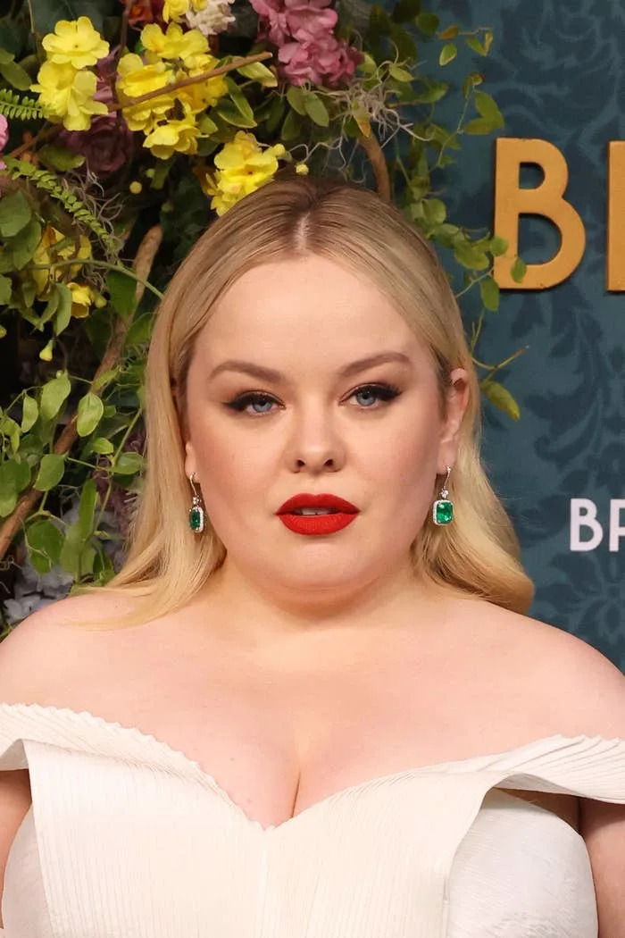 Nicola Coughlan wearing an off-the-shoulder dress with statement earrings stands in front of a floral backdrop at a public event