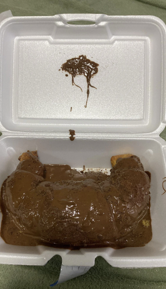 A Styrofoam container holds a croissant covered in chocolate sauce, with some sauce smeared on the container lid