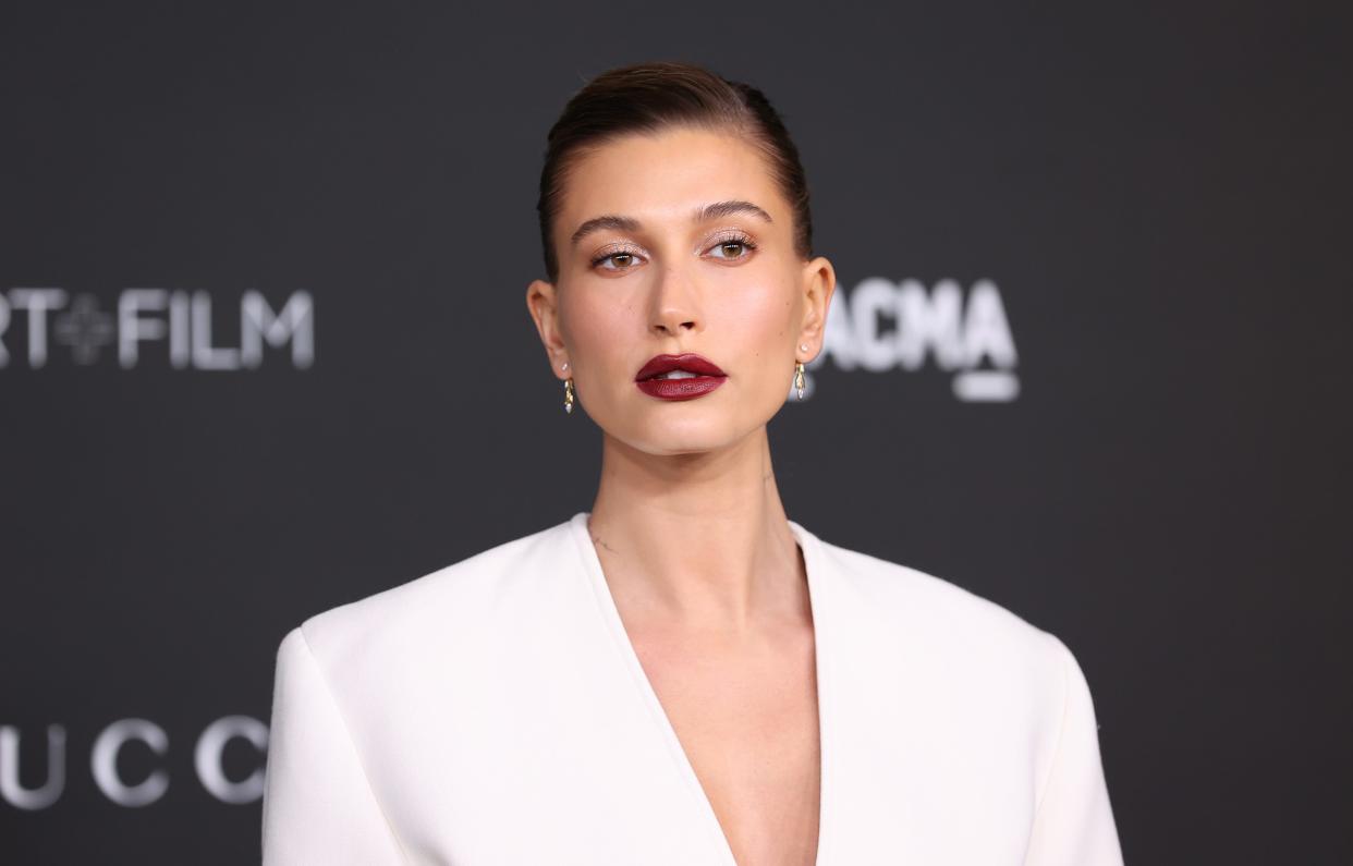 Hailey Bieber at an event in a white dress and dark red lipstick.