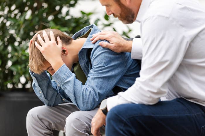 A man in a white shirt comforts a distressed young boy in a denim jacket, who is sitting with his head in his hands