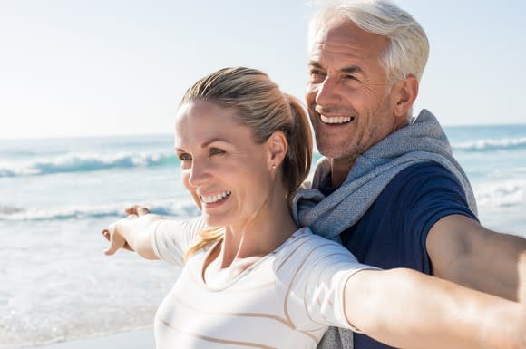 Older couple smiling, standing on a beach.