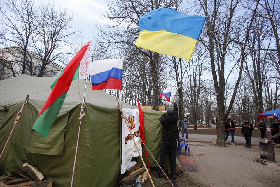 In this photo taken Tuesday, March 11, 2014, a man places a Ukrainian flag atop a tent alongside flags from Russia, center, and Belarus, left, at a tent camp set up by pro Russia activists in Luhansk, eastern Ukraine. Luhansk was home to one of the Soviet Union’s blue-ribbon factories that turned out locomotives deemed good enough to be designated IS--the Russian-language initials of Josef Stalin. Since Russian troops rolled into Crimea, and lawmakers there scheduled a referendum for Sunday on whether to join Russia, the world’s attention has focused on the fate of the lush peninsula that juts into the Black Sea. But here in Ukraine’s coal-fired industrial east, where Russians have lived for more than two centuries, a potent mix of economic depression, ethnic solidarity and nostalgia for the certainties of the Soviet past have many demanding the right to become part of Russia as well.(AP Photo/Sergei Grits)