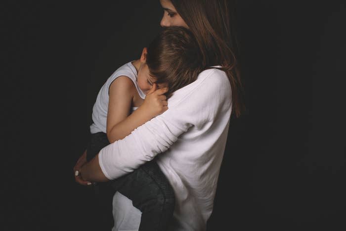 A woman is holding a young boy closely, both are facing sideways. The woman is wearing a long-sleeved top, and the boy has a sleeveless shirt