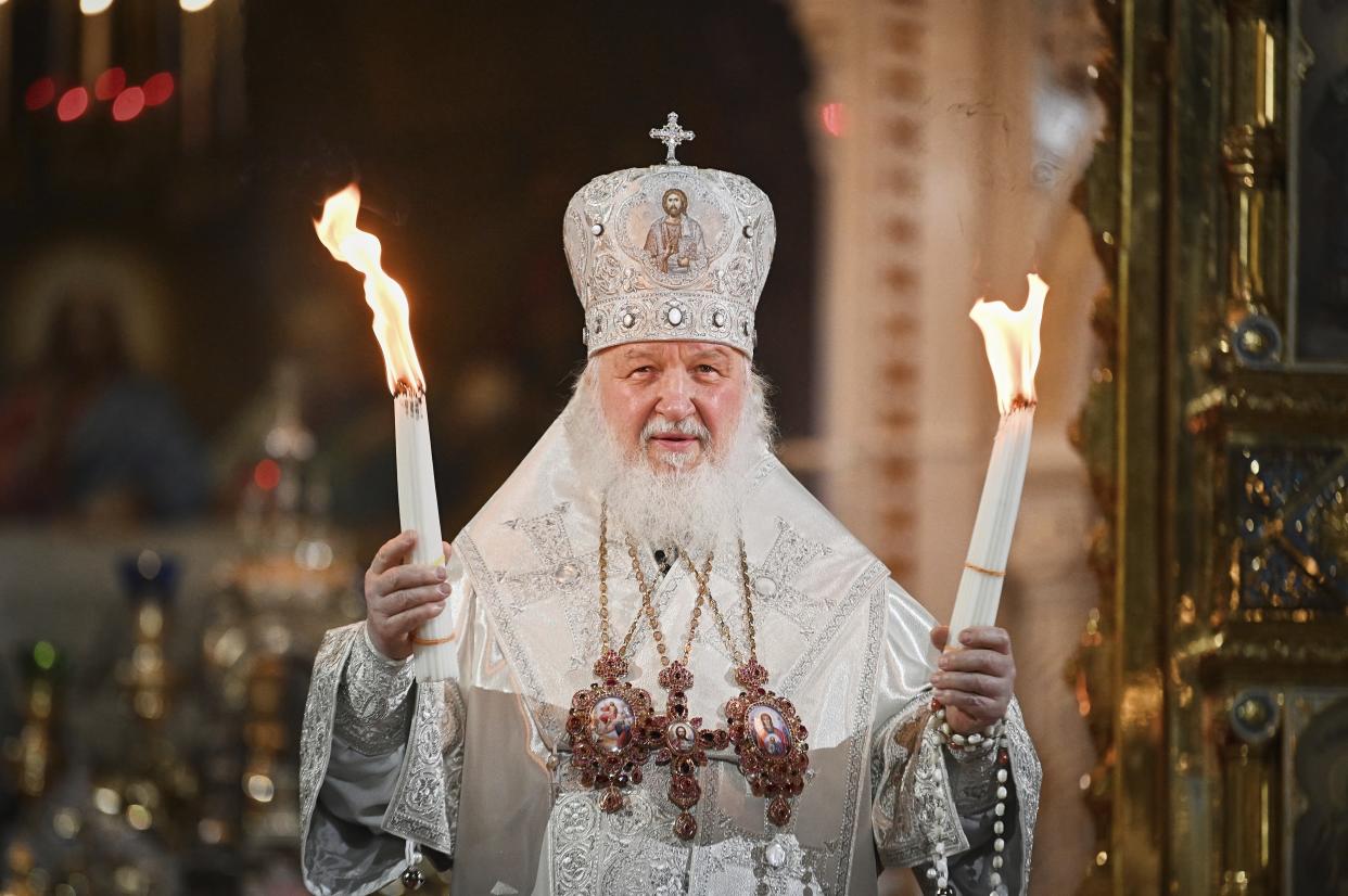 Patriarch Kirill, in white jeweled miter, white robes and with three icons suspended from his neck, holds a candle in each hand by an ornate brass gate to the altar..