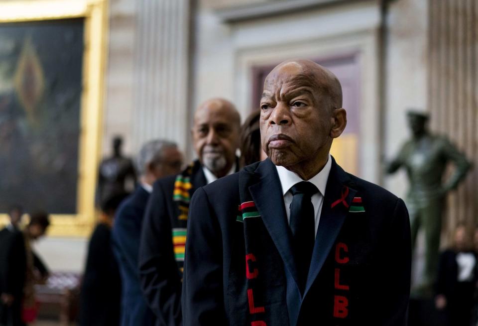 john lewis stands inside a large room in a blue suit jacket and tie, he also wears a black scarf with red letters and a red and green stripe, he looks to the left and people stand behind