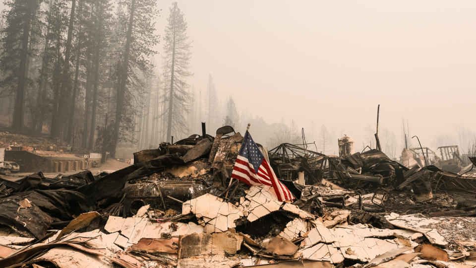 An American flag rests on a pile of burned rubble, with trees seen in the background through haze.