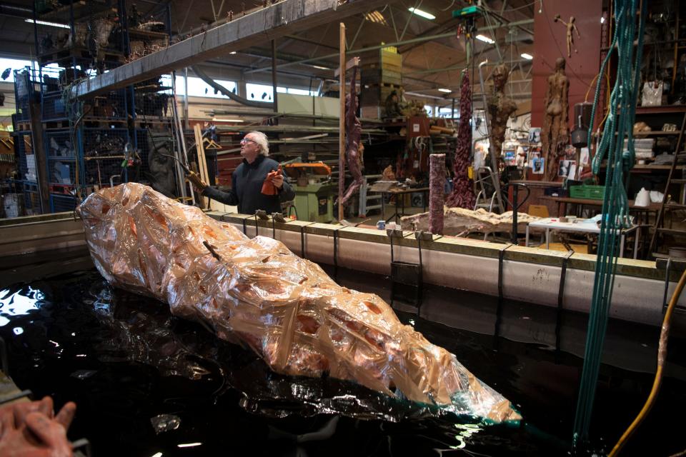 A sculpture is seen wrapped in a cramped artists studio, and a man is seen pressing a remote in the background of the image.