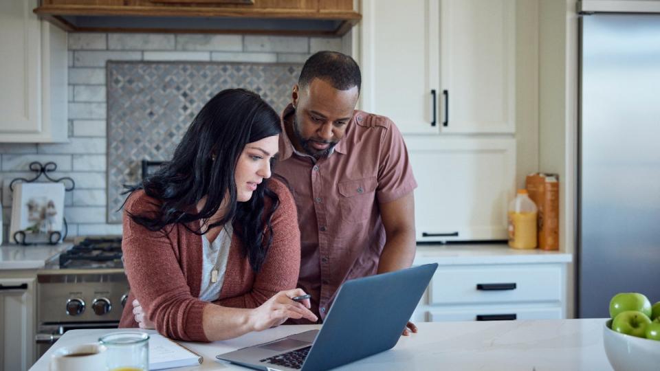 Caucasian woman and African American man sit at kitchen counter with breakfast working with pen, paper and laptop