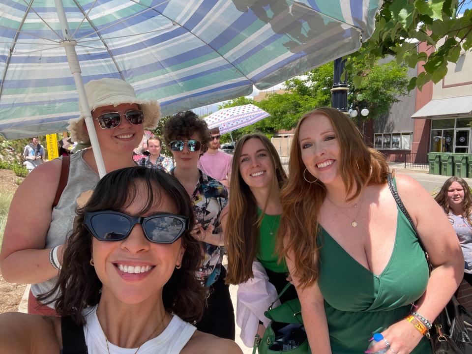 The writer with sunglasses on smiling with other individuals in line under an umbrella