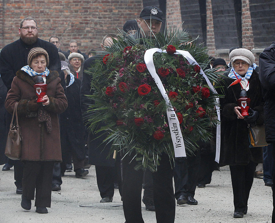 Two women, Auschwitz survivors, flank a huge floral wreath of red roses and palm fronds, with a brick wall behind the crowd of attendees. 