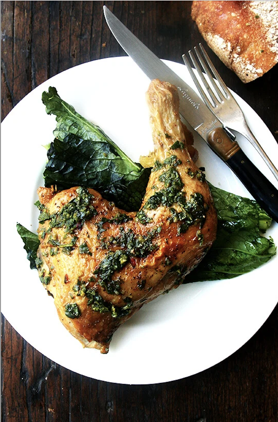 A roasted chicken leg garnished with herbs, served on a bed of greens with a knife and fork on a white plate. A piece of bread is partially visible