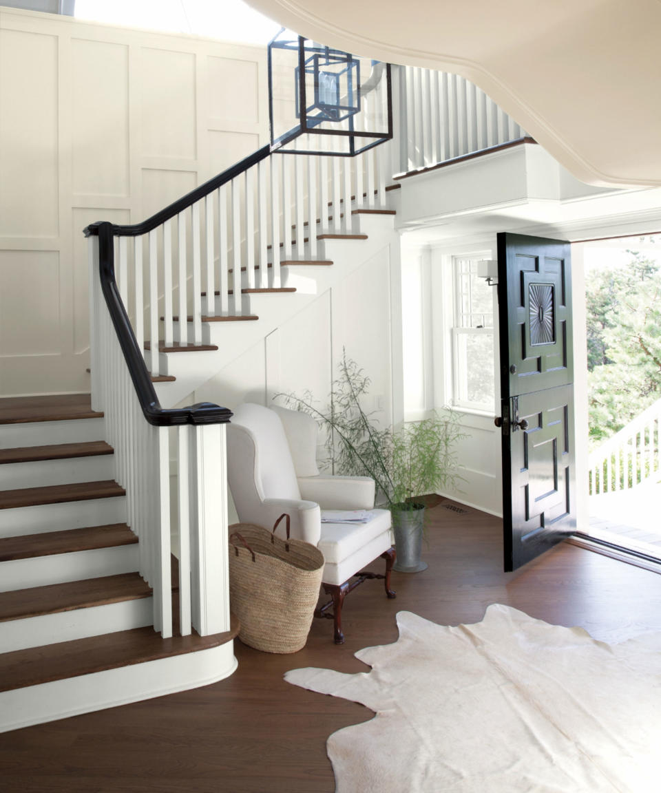 Large hallway with white painted walls and dark wood flooring, unique geometric design on black front door, animal skin style rug, white paneling on walls, cream lounge chair and plant, black painted handrail and pendant light