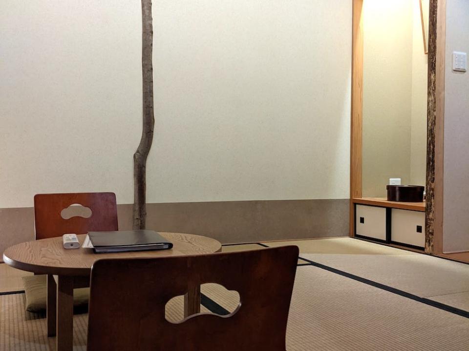 A photo of two wooden chairs without legs resting on the tatami mat floor around a small wooden table. Opposite, in the right corner of the room is a small alcove with a shelf that has a wooden box and below that two sliding cupboard doors. The walls are white with a thin carved wooden tree running from ceiling to floor. The ceiling is made with bamboo beams.