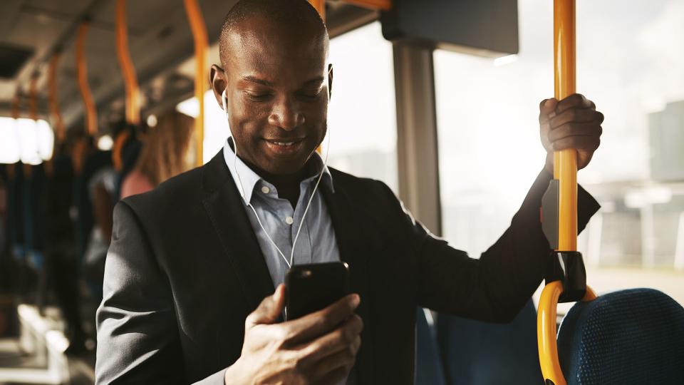Smiling young African businessman wearing a suit standing on a bus during his morning commute listening to music on a smartphone and earphones.