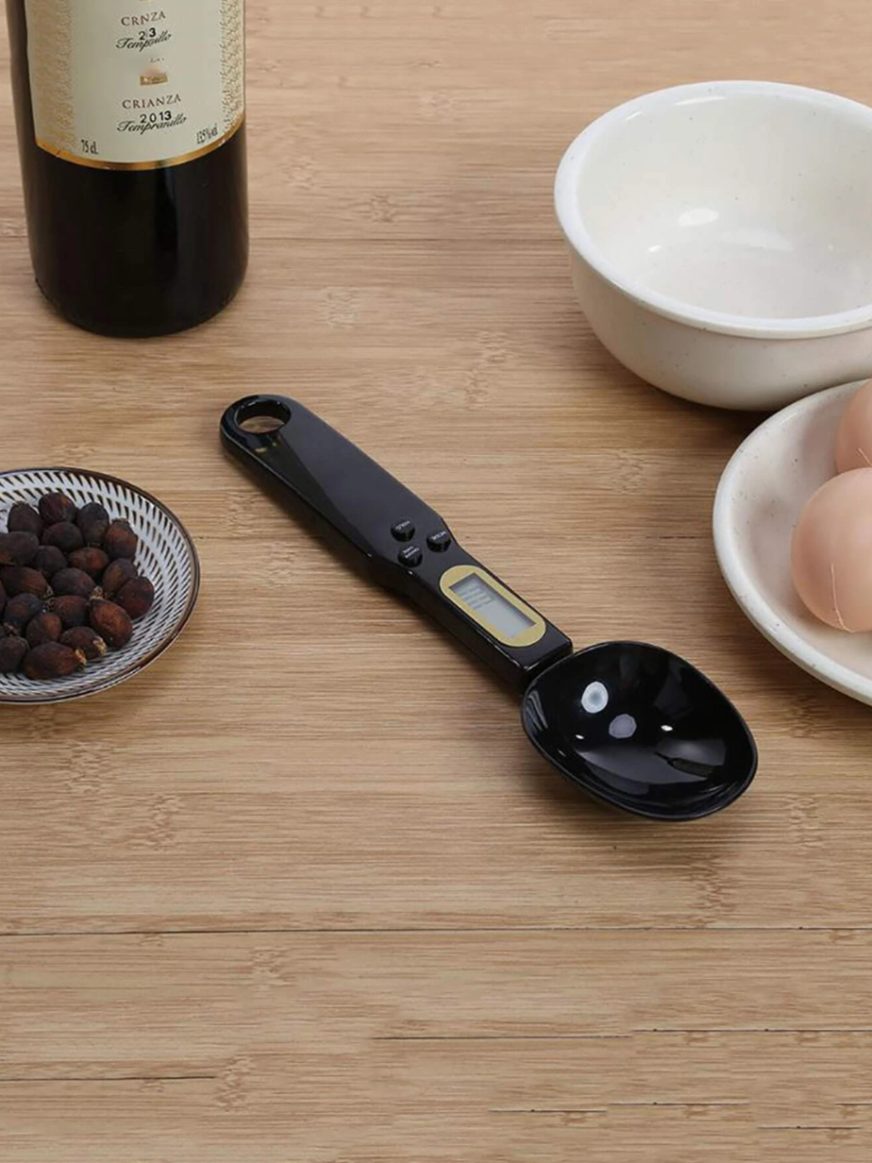 Black electronic measuring spoon is pictured on wooden kitchen bench with bowls, eggs and kitchen ingredients.