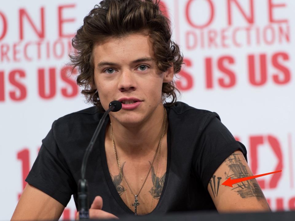 A red arrow pointing to a tattoo of three nails on Harry Styles' left bicep.