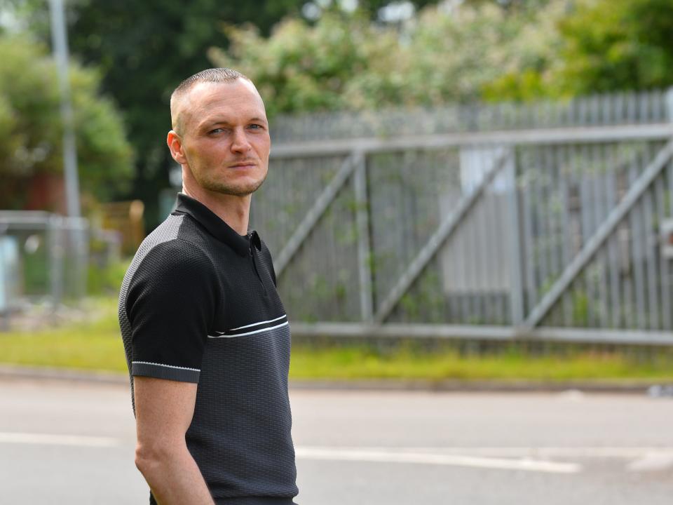 A man stands looking at the camera in front of a road and a metal fence.