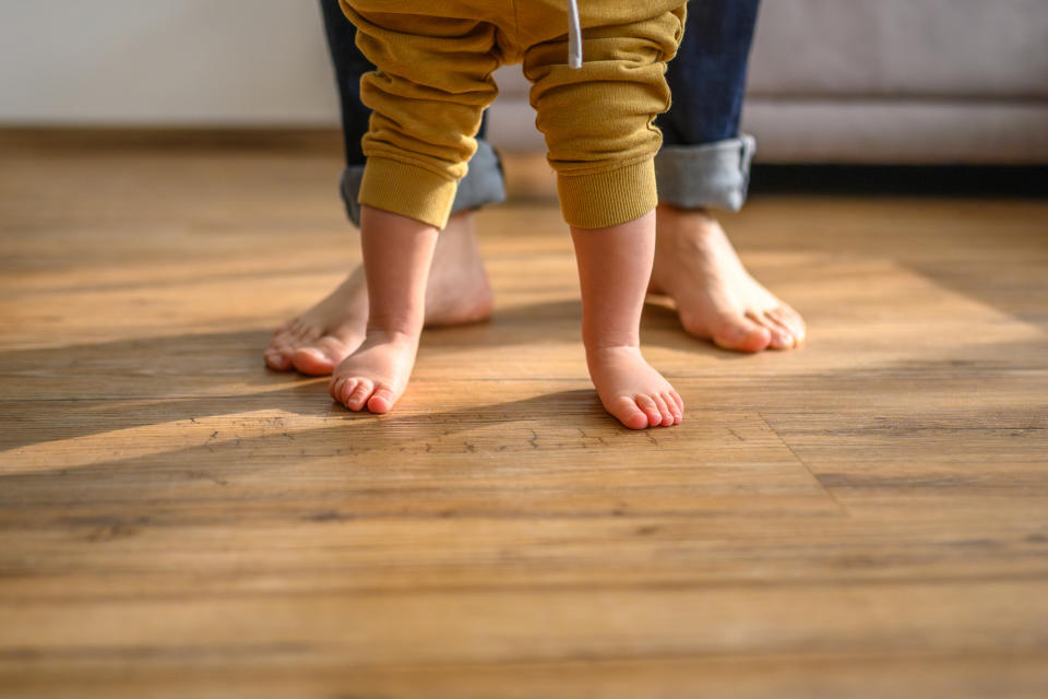 Two sets of bare feet stand on a wooden floor. An adult stands behind a toddler who is taking steps