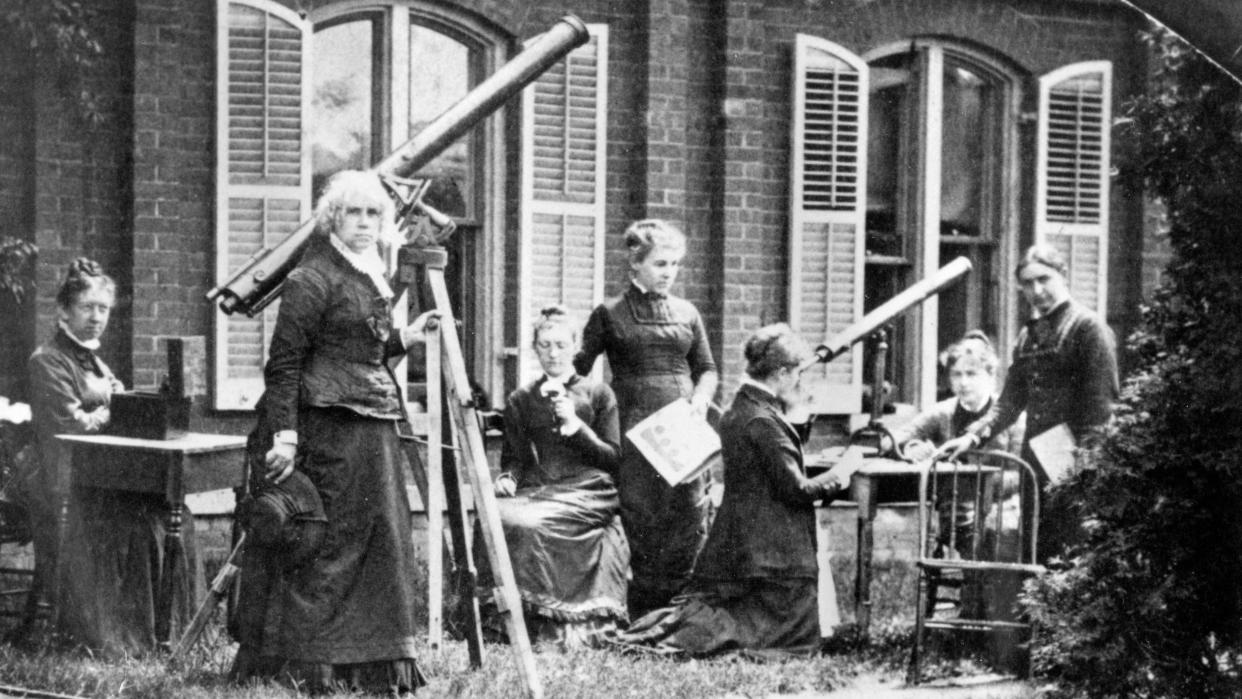  photograph of several women in the 1870s. the women all wear black dresses. some are sitting at tables. some are standing. maria mitchell is near the middle of the picture beside a long telescope. behind is a building with window shades open 