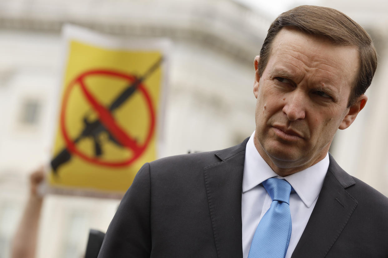 Chris Murphy, in a suit jacket and blue tie, in front of a sign with a picture of a gun with a slash through it.