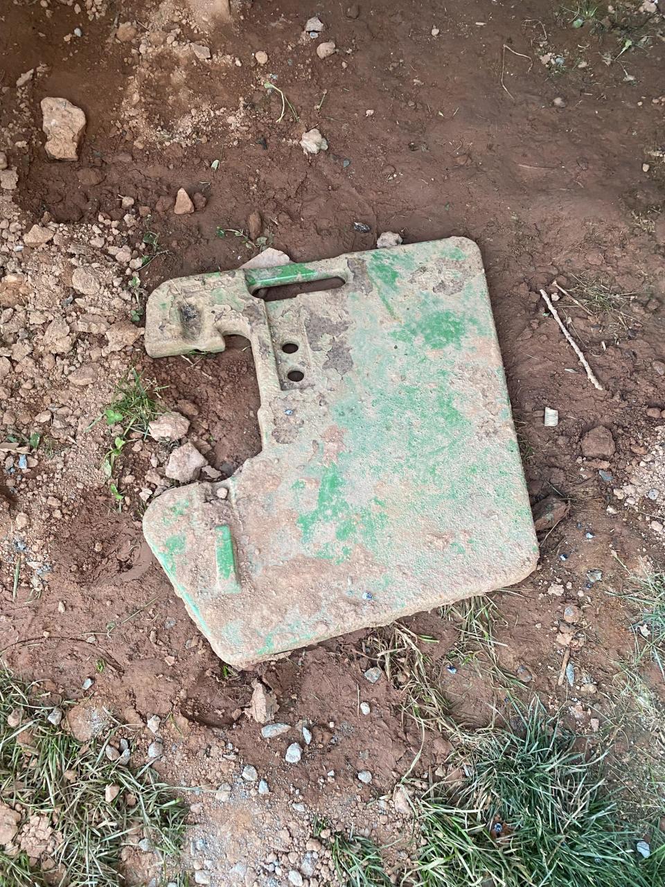 A worn out rectangular piece of equipment with holes, screw slots and dirt stains lies on the ground surrounded by dirt and grass