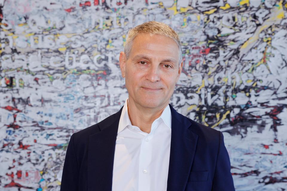 ariel emanual, ceo of endeavor, smiles at the camera wearing a white button down, navy suit jacket, standing in front of a colorful white background