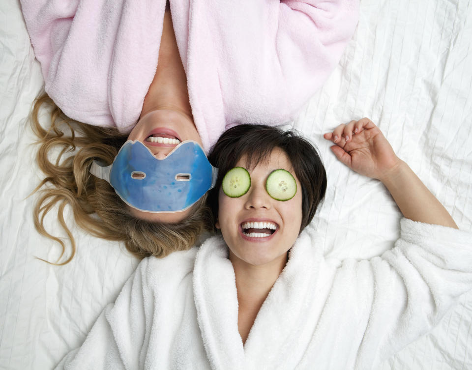 Two women lying on a bed in bathrobes, one with an eye mask on and the other with cucumbers over her eyes. 