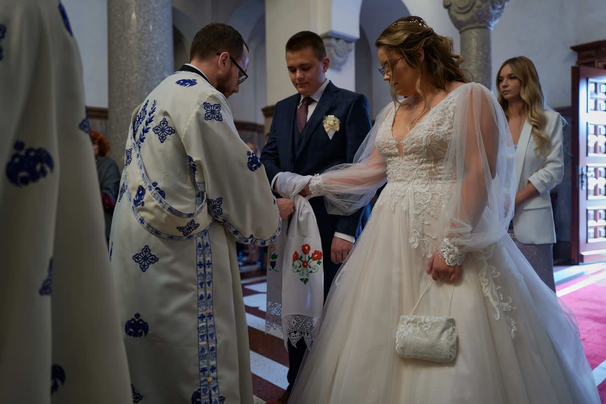 A priest binds the hands of a person wearing a white dress and another person wearing a blue suit. 