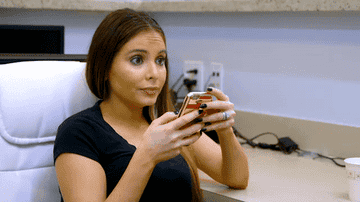 A woman with long brown hair, wearing a black shirt, sits in a white chair looking at her phone with a surprised expression