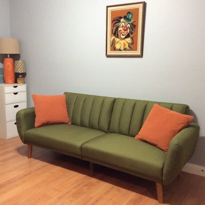 A green mid-century modern sofa with two orange pillows, under a framed painting of Goofy, in a room with home decor