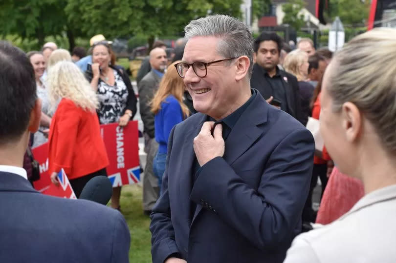 Sir Keir Starmer pictured in black suit and shirt adjusting his collar and smiling