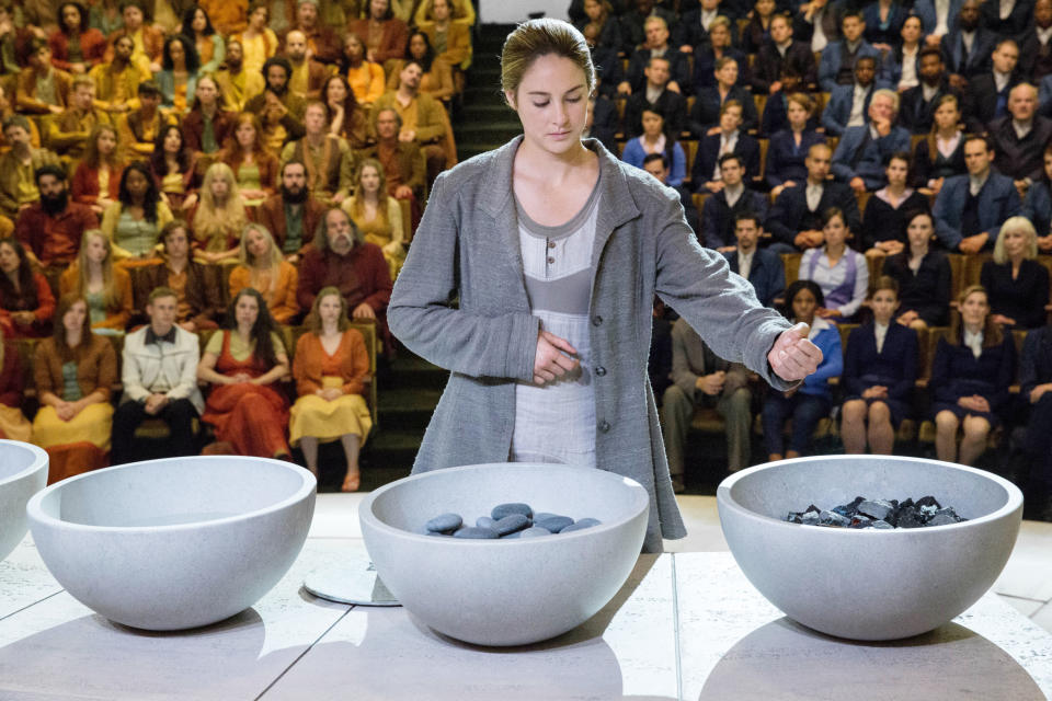 Shailene Woodley in a scene from "Divergent," surrounded by large bowls of stones, in front of a seated audience. She is wearing a loose jacket and a light dress
