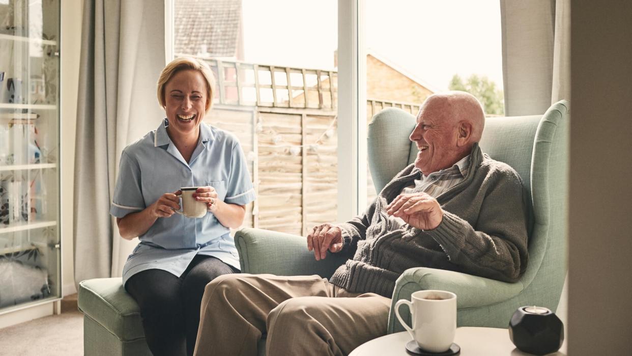 Indoor shot of smiling senior man and female carer enjoying coffee in living room.